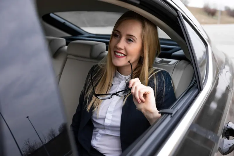 A sophisticated businesswoman sits in the back seat of a luxury black sedan, smiling and holding her glasses, representing an elegant IAH to Galveston Port transfer experience.